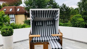 A sun lounger with a striped cushion and a wooden frame, positioned on a tiled patio with a potted plant nearby.