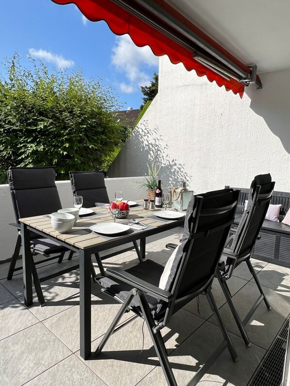 Un patio al aire libre con una mesa dispuesta para dos personas, rodeado de sillas negras, un toldo rojo y una pared blanca.