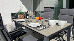 An outdoor dining area with a table set for two, including plates, cutlery, glasses, and a bottle of wine. There are two chairs with cushions, a potted plant, and a window with a view of the sky and a building.