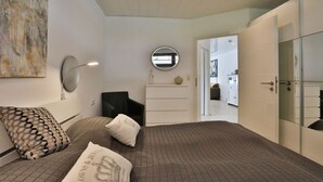 A modern bedroom with a bed, a dresser, a mirror, and a door leading to another room.