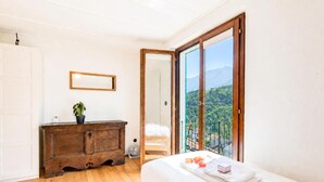 A bedroom with a wooden dresser, a bed with neatly folded towels, a potted plant, and a large sliding glass door leading to a balcony with a mountain view. - La Canonica Centovalli with Balconies and Garden, Camedo, Switzerland (Camedo)