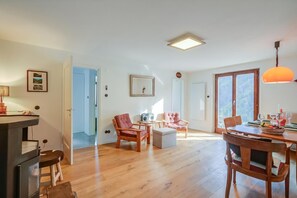 Ein geräumiges Zimmer mit Holzboden, einem Esstisch für vier Personen, zwei Sesseln, einem kleinen Tischchen mit Radio und einem großen Fenster mit Blick.