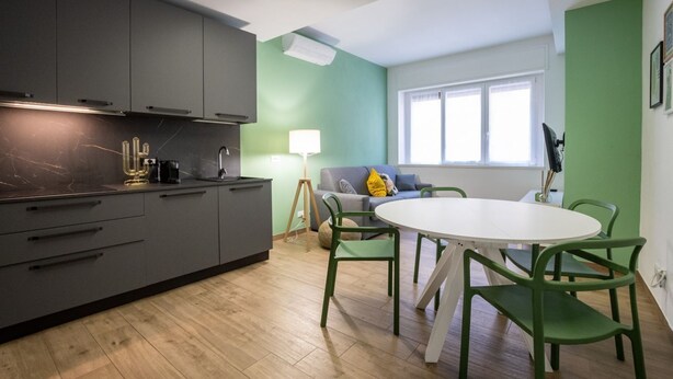 A modern kitchen with grey cabinets, a marble countertop, and a coffee machine. There is a dining area with a round table and green chairs. The room has a green accent wall and a window with white curtains. - Gorki 11 Modern Escape (Milano)