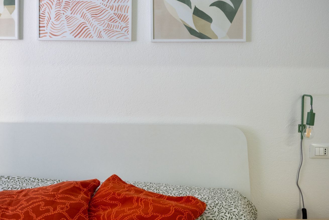 Ein Bett mit gemusterten Textilien, eine Steckdose mit grünem Stecker und gerahmte Kunstwerke an der Wand.