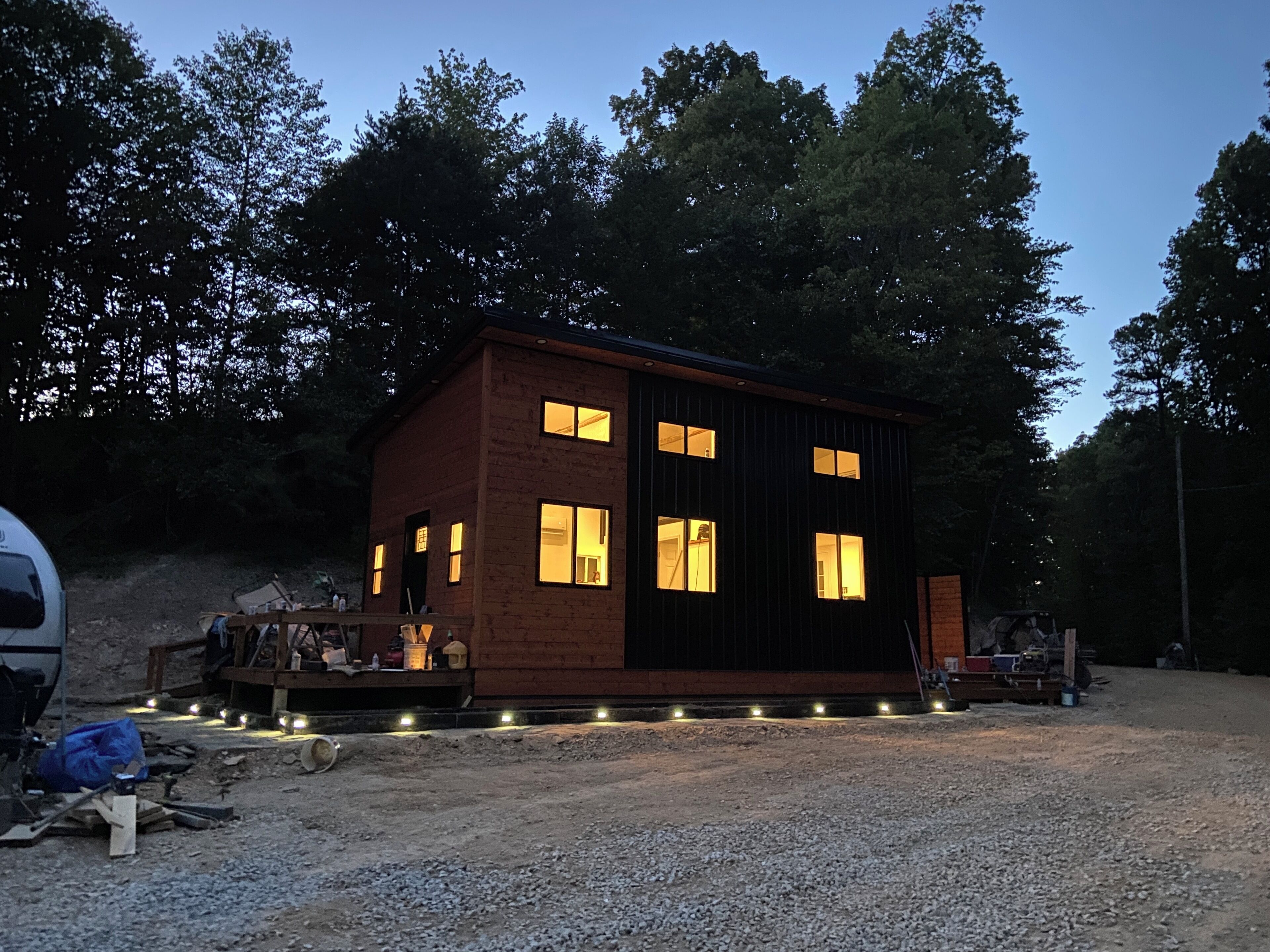 A small, two-story cabin with multiple windows, surrounded by trees and gravel ground.