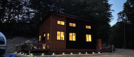 A small, two-story cabin with multiple windows, surrounded by trees and gravel ground.