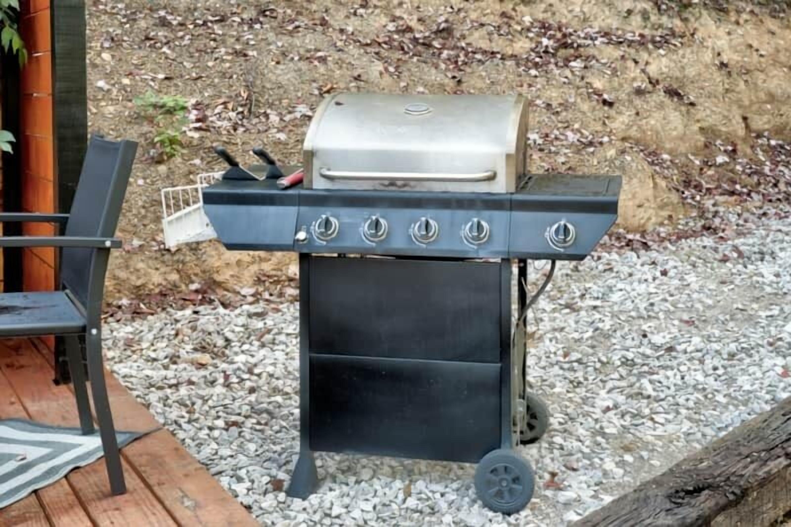 A portable grill with four burners and a side shelf, placed on a gravel surface with a wooden deck and a chair nearby.