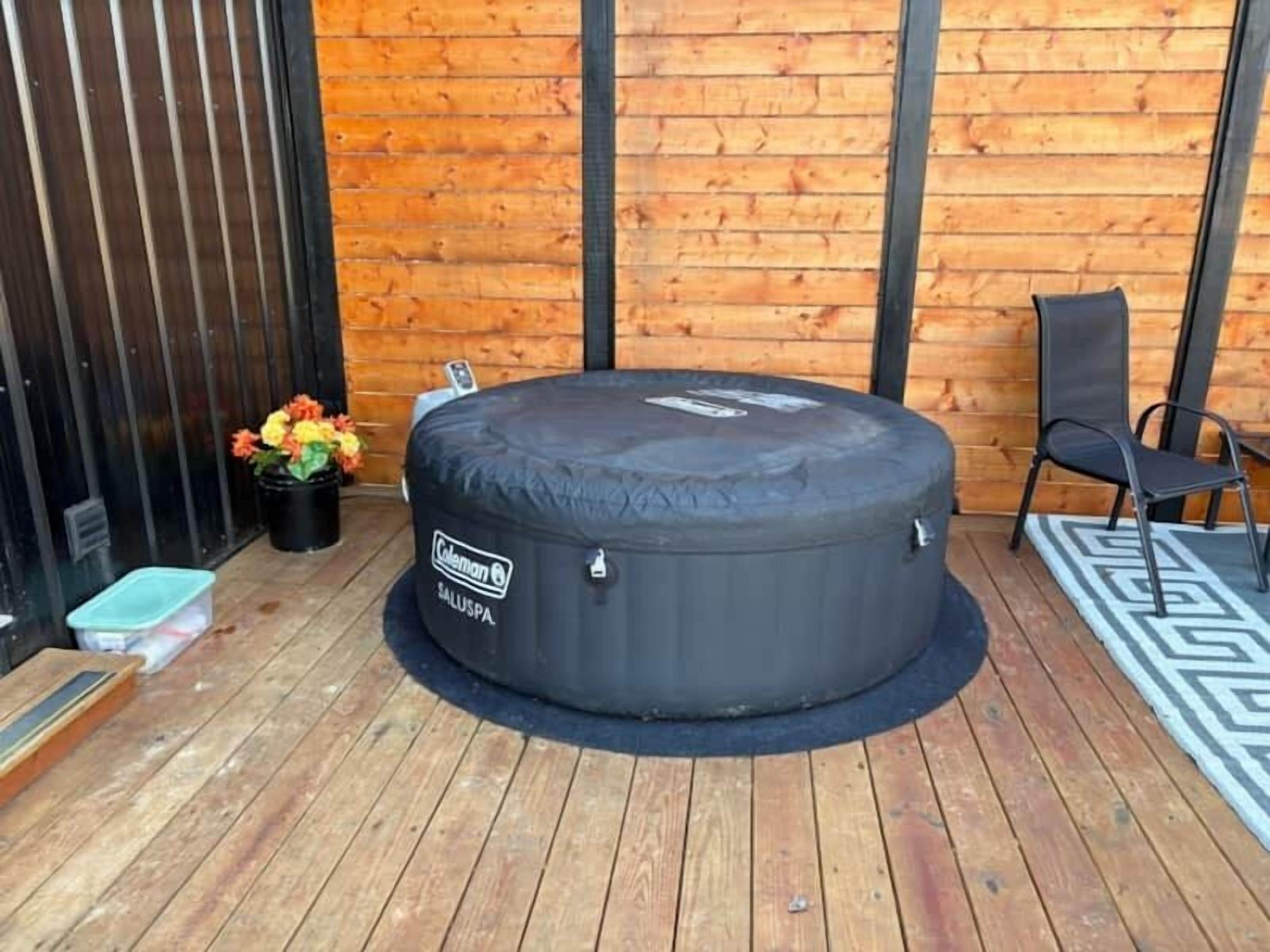 A round, black outdoor seating area with a Coleman brand logo, a wooden deck, a black chair, and a potted plant with flowers.
