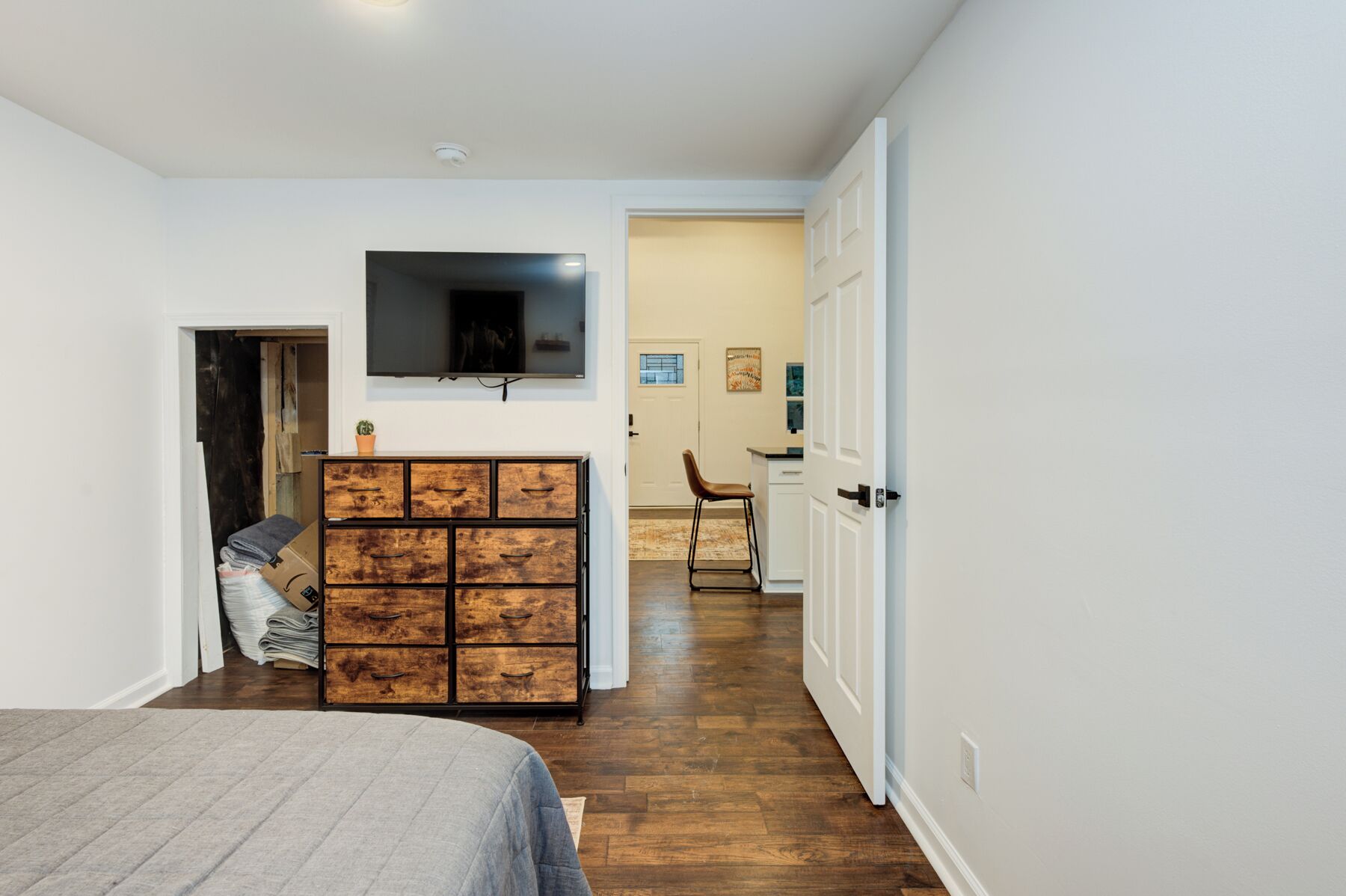 A bedroom with a bed, a wooden chest of drawers, a television mounted on the wall, and a door leading to another room.