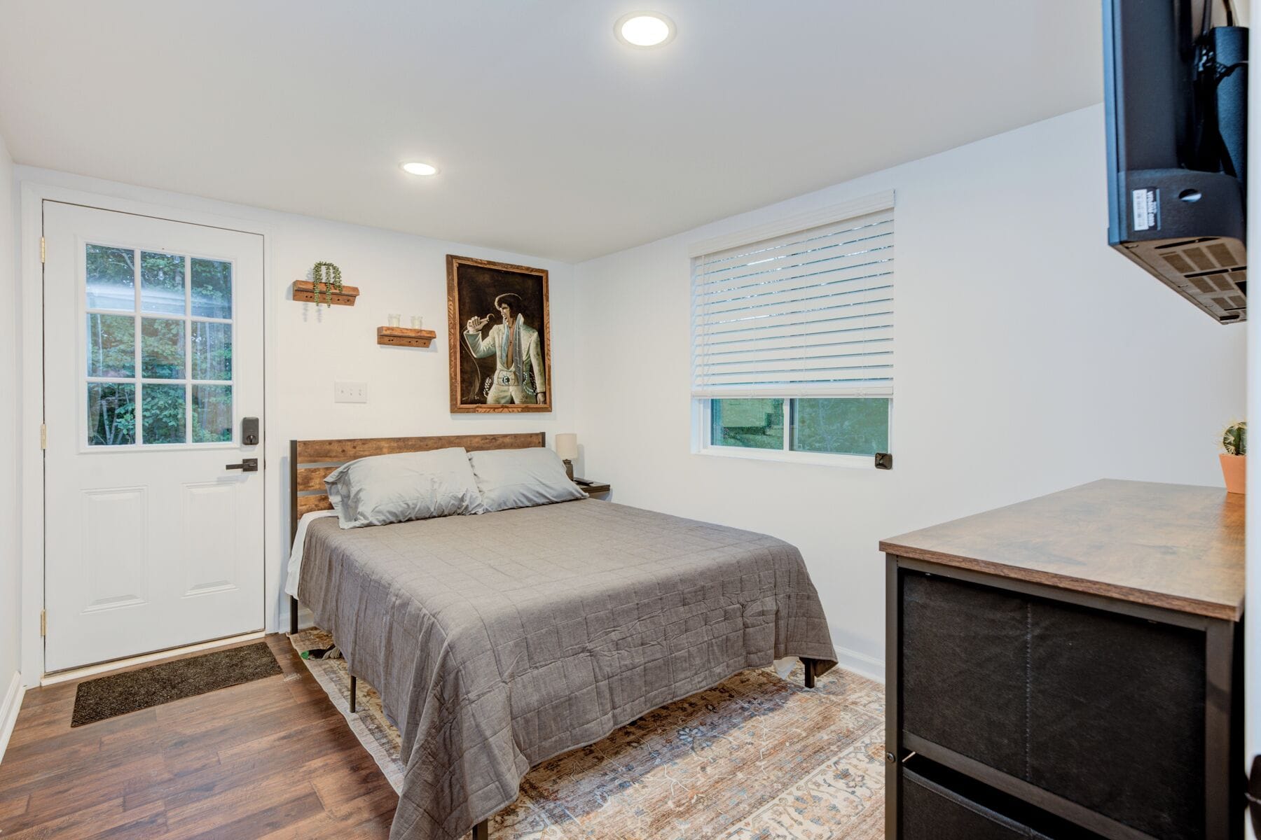 A bedroom with a bed, a bedside table, a window with blinds, a door, and a small kitchenette area.