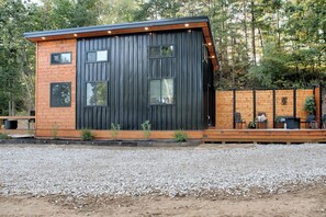 A modern cabin with a wooden deck and a gravel driveway.