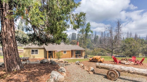 A rustic cabin with a porch, surrounded by a gravel area with rocks and logs.