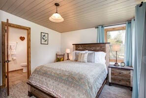 Un dormitorio con cabecera de madera, colcha estampada, mesita de noche con lámpara y ventana con cortinas.