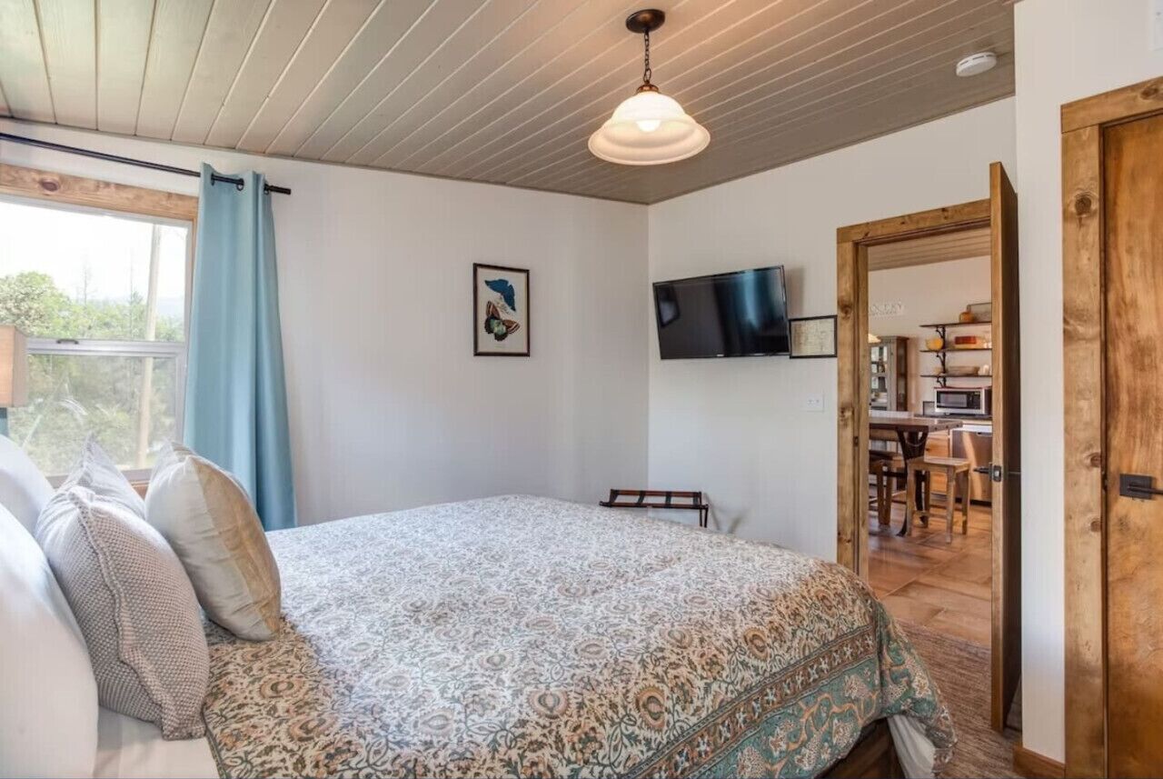 Un dormitorio con una colcha estampada, un televisor montado y una puerta de madera que da a una zona de cocina.