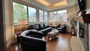 A modern living room with a black leather sofa, a wooden coffee table, and a fireplace. Large windows offer a view of the cityscape. - Solara Corner Suite-Pool & Hot Tub & Spa (Canmore)