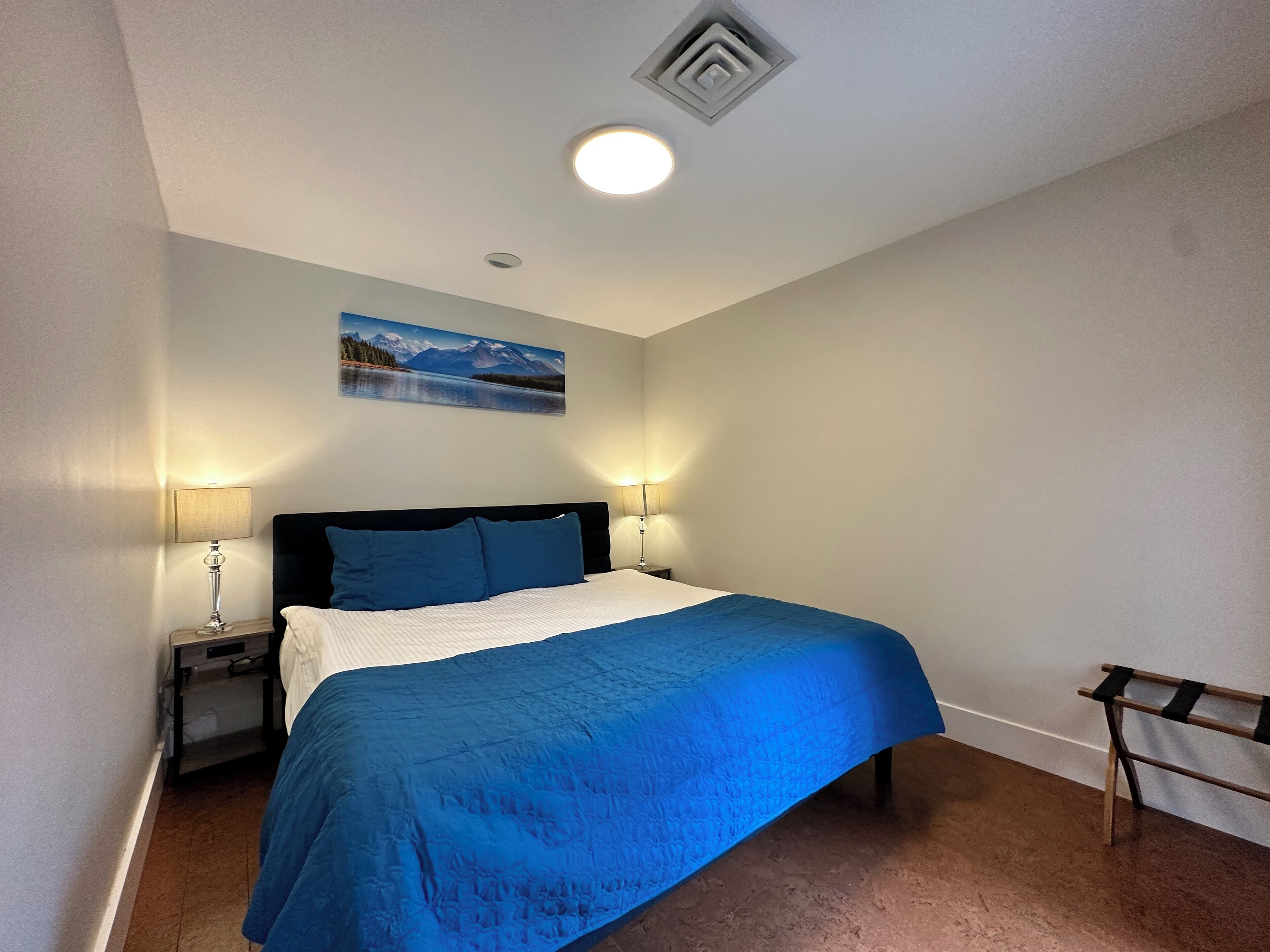 A bedroom with a bed, two bedside lamps, a nightstand, a painting on the wall, and a wooden bench.