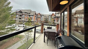A balcony with wicker furniture, a view of multi-story buildings, and parked cars. - Solara Corner Suite-Pool & Hot Tub & Spa (Canmore)