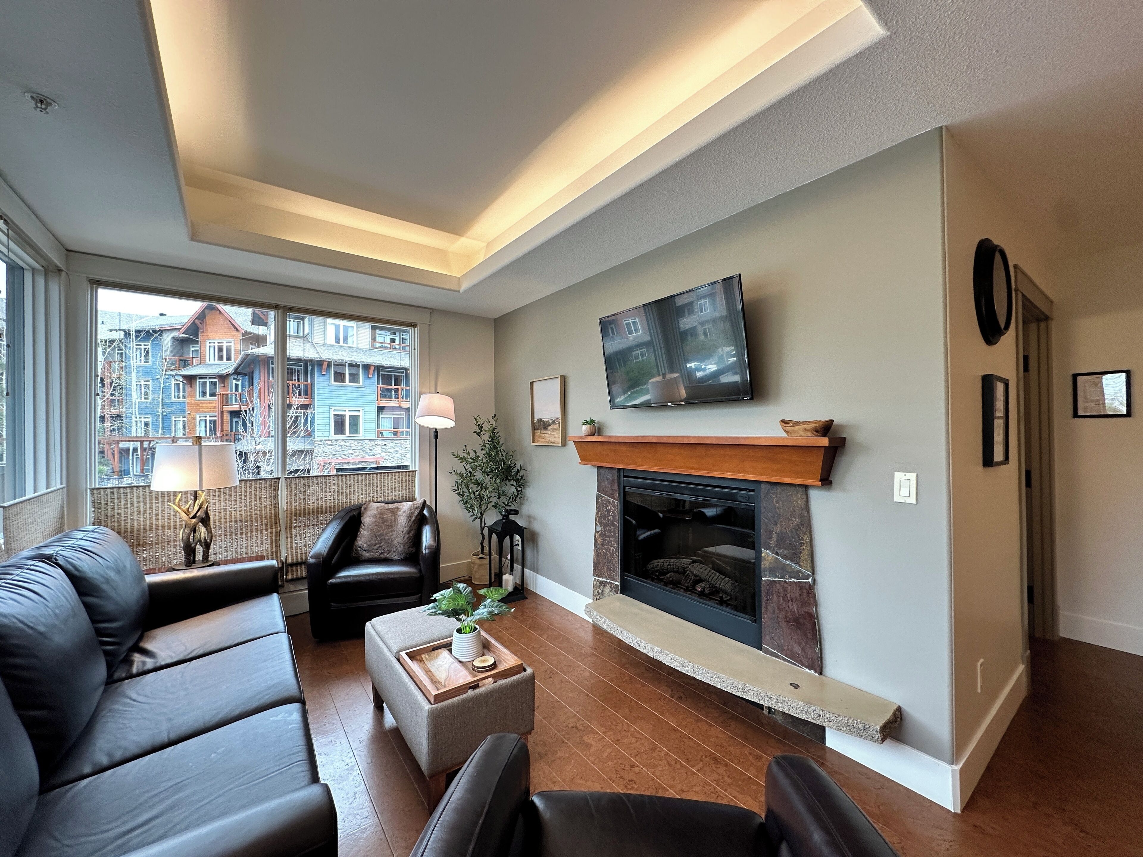 A modern living room with a fireplace, comfortable seating, a flat-screen TV, and a view of a residential area.