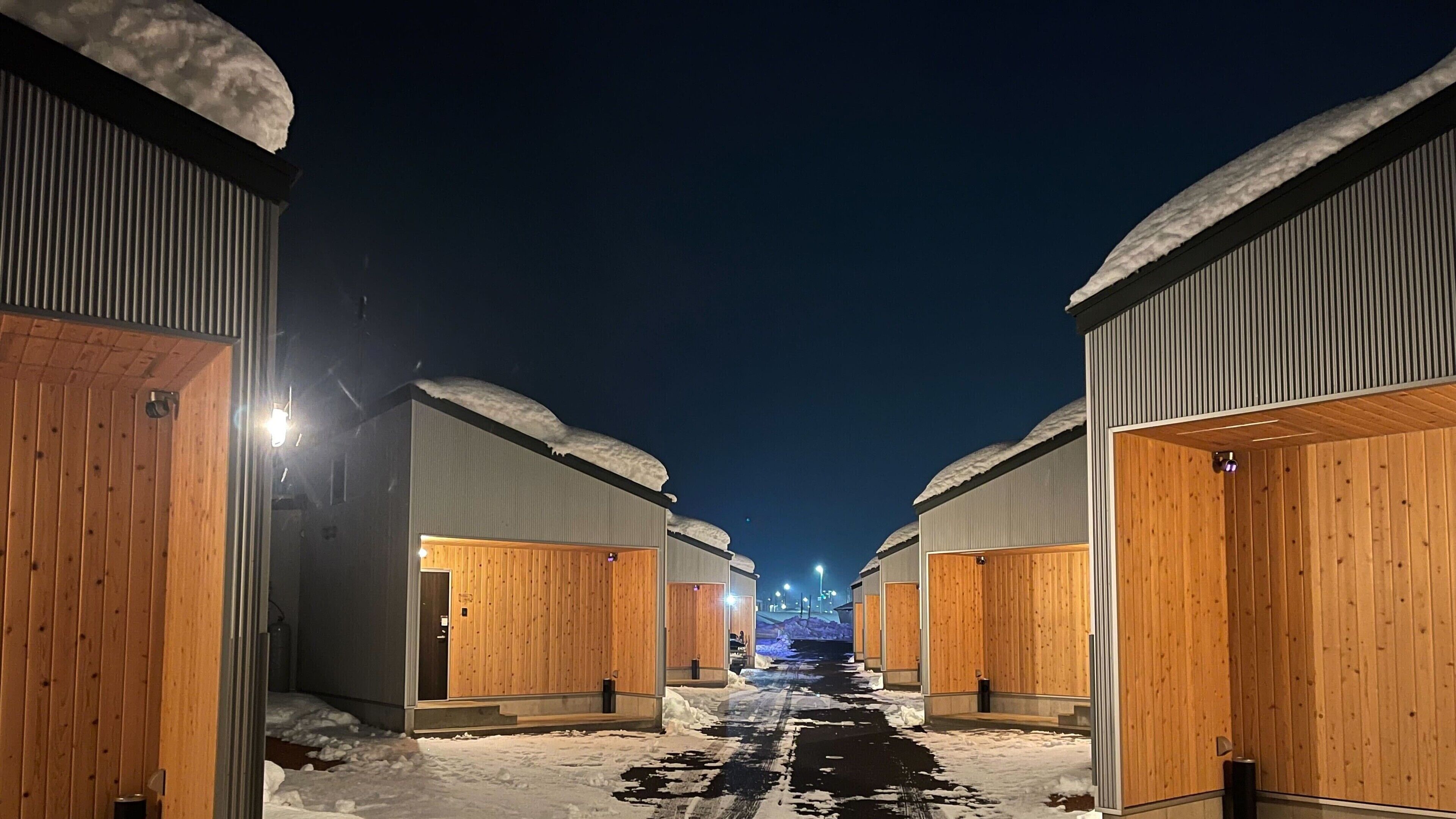 A snow-covered pathway leading to a row of wooden cabins with metal exteriors, each with a door and a small window.