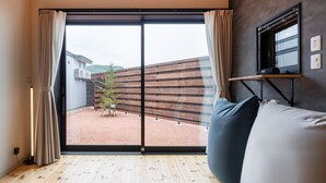 A room with a sliding glass door leading to a covered outdoor area, a wooden floor, a grey sofa, and a mounted TV on the wall. - HOTEL SOBOKU (Myoko)