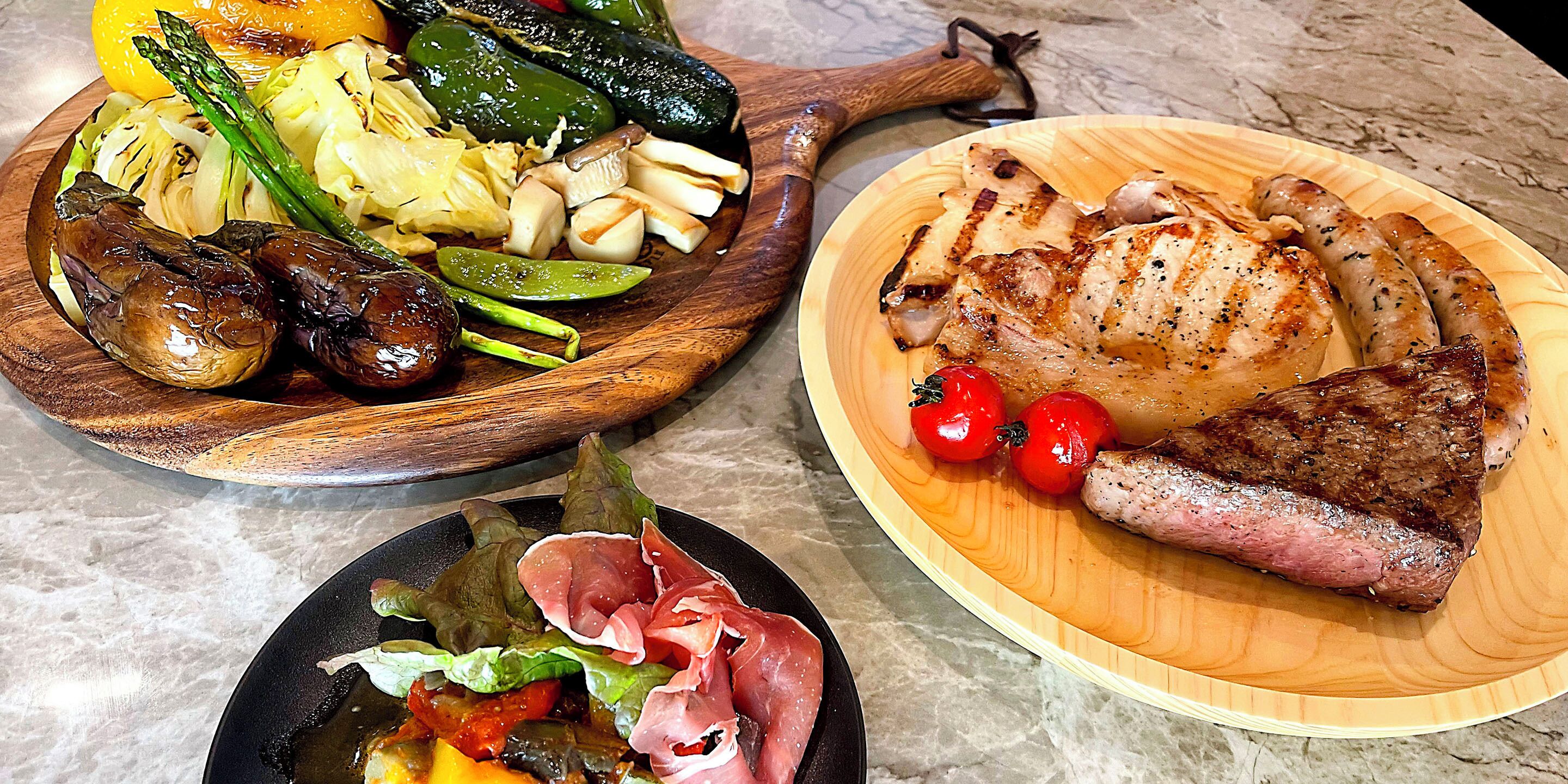 A variety of dishes including grilled meats, vegetables, and sausages on wooden serving platters.