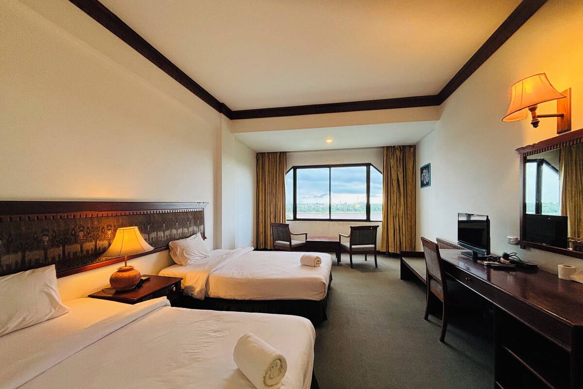 A hotel room with two beds, a desk with a computer, a chair, and a large window offering a view.