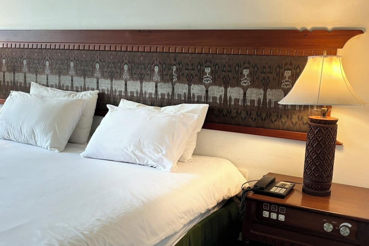 A hotel room with a large bed, white linens, a headboard with a patterned design, a bedside lamp, and a nightstand with a phone and a remote control.