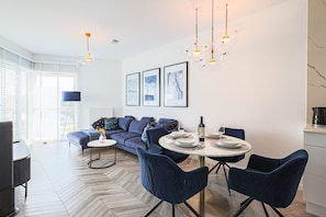 A modern living room with a sofa, dining table, and chairs, a large window, and a chandelier. - 2Homerent Piedra | Relax with a Baltic Sea View (Gąski)