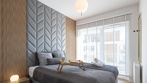 A modern bedroom with a large bed, a bedside table with a lamp, a tray with a cup and saucer, and a window with blinds. - 2Homerent Piedra | Relax with a Baltic Sea View (Gąski)