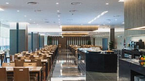 Un comedor moderno de hotel con mesas y sillas largas, un mostrador de buffet con comida y bebidas, y un interior limpio y bien iluminado.