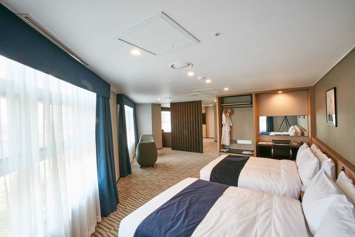 A hotel room with two beds, a large window with curtains, a mirror, and a seating area.