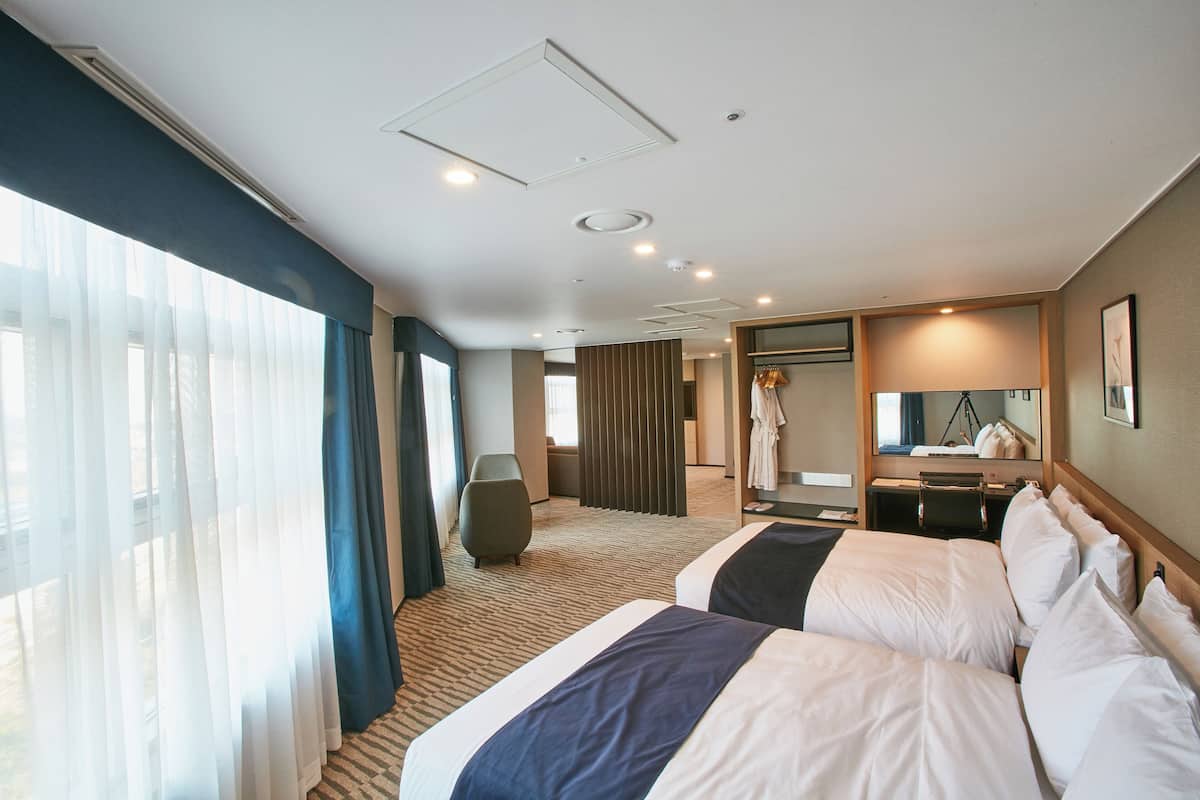 A hotel room with two beds, a large window with curtains, a mirror, and a seating area.