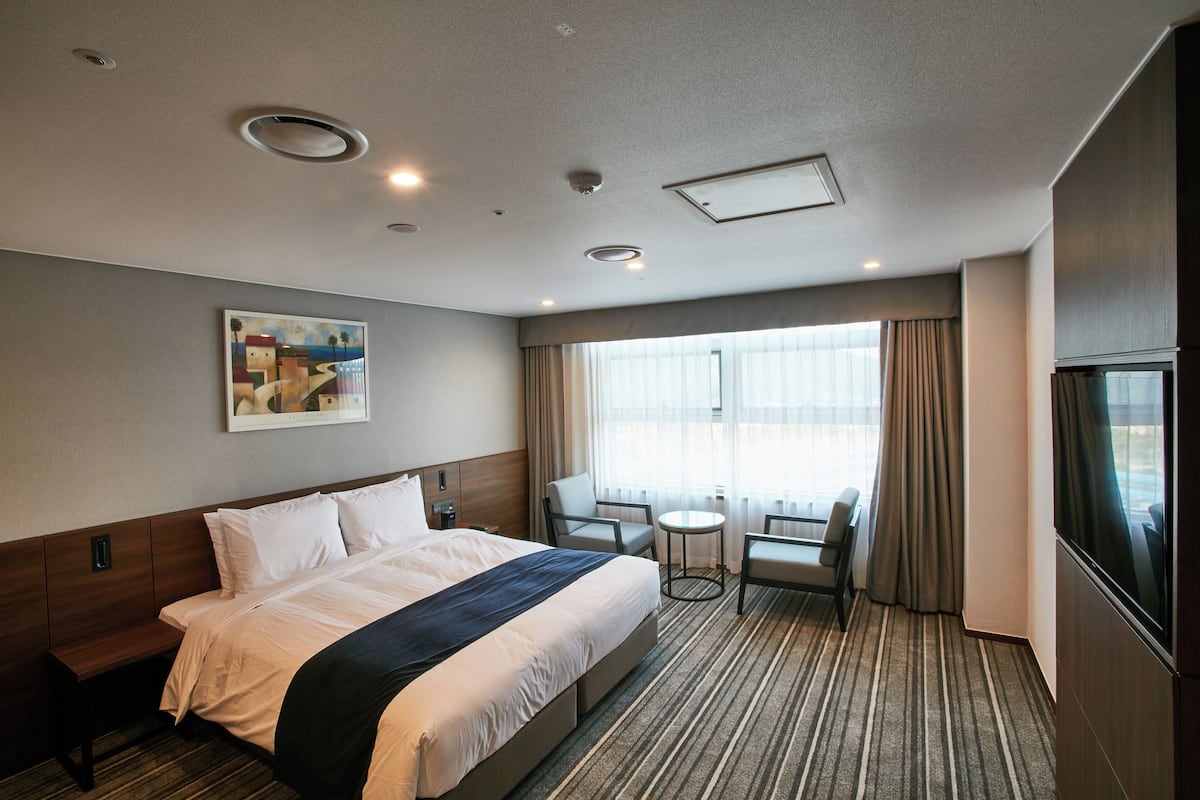 A hotel room with a bed, a small table, two chairs, a window with curtains, and a painting on the wall.