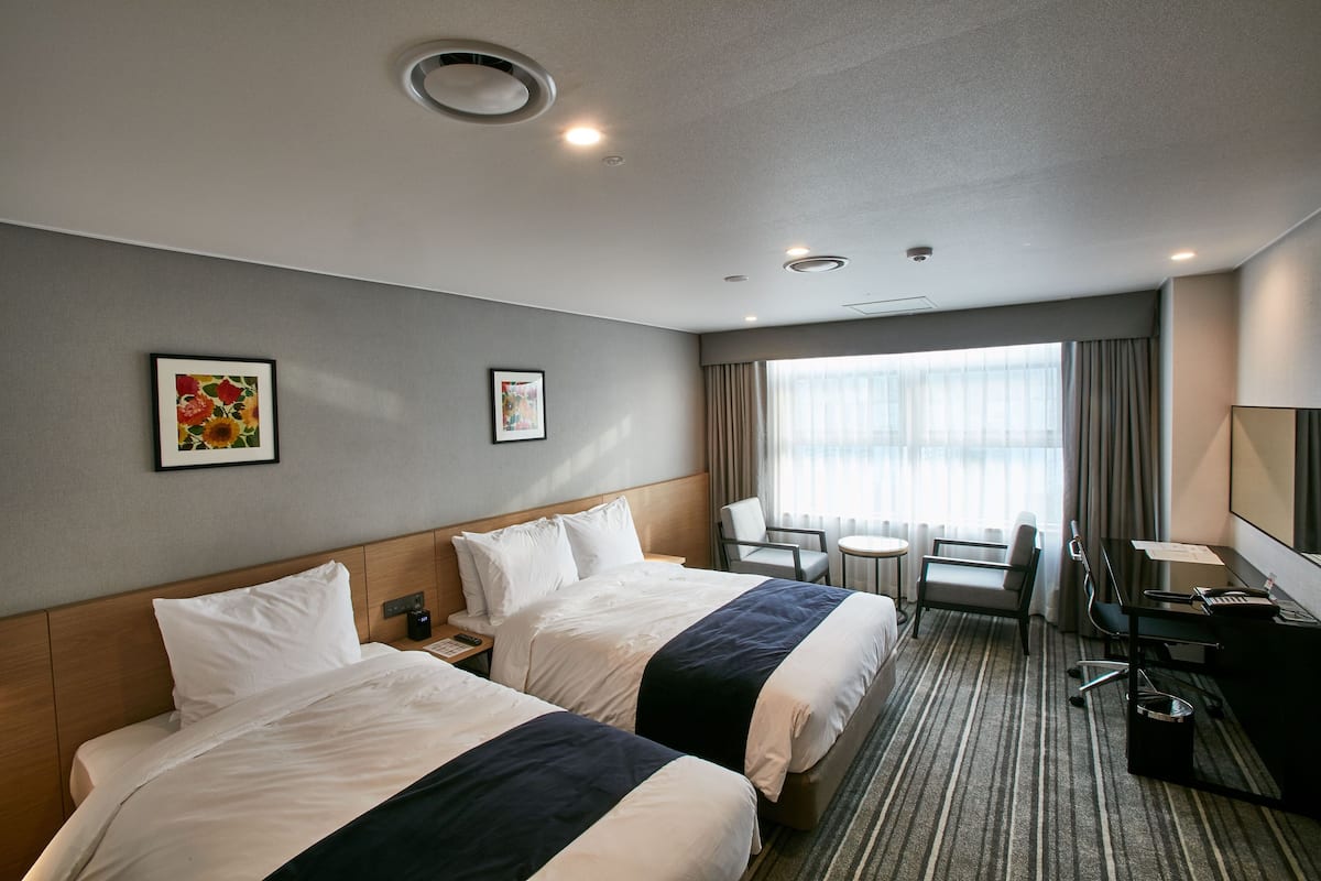 A hotel room with two beds, a desk, a chair, a small table, and a large window with curtains.