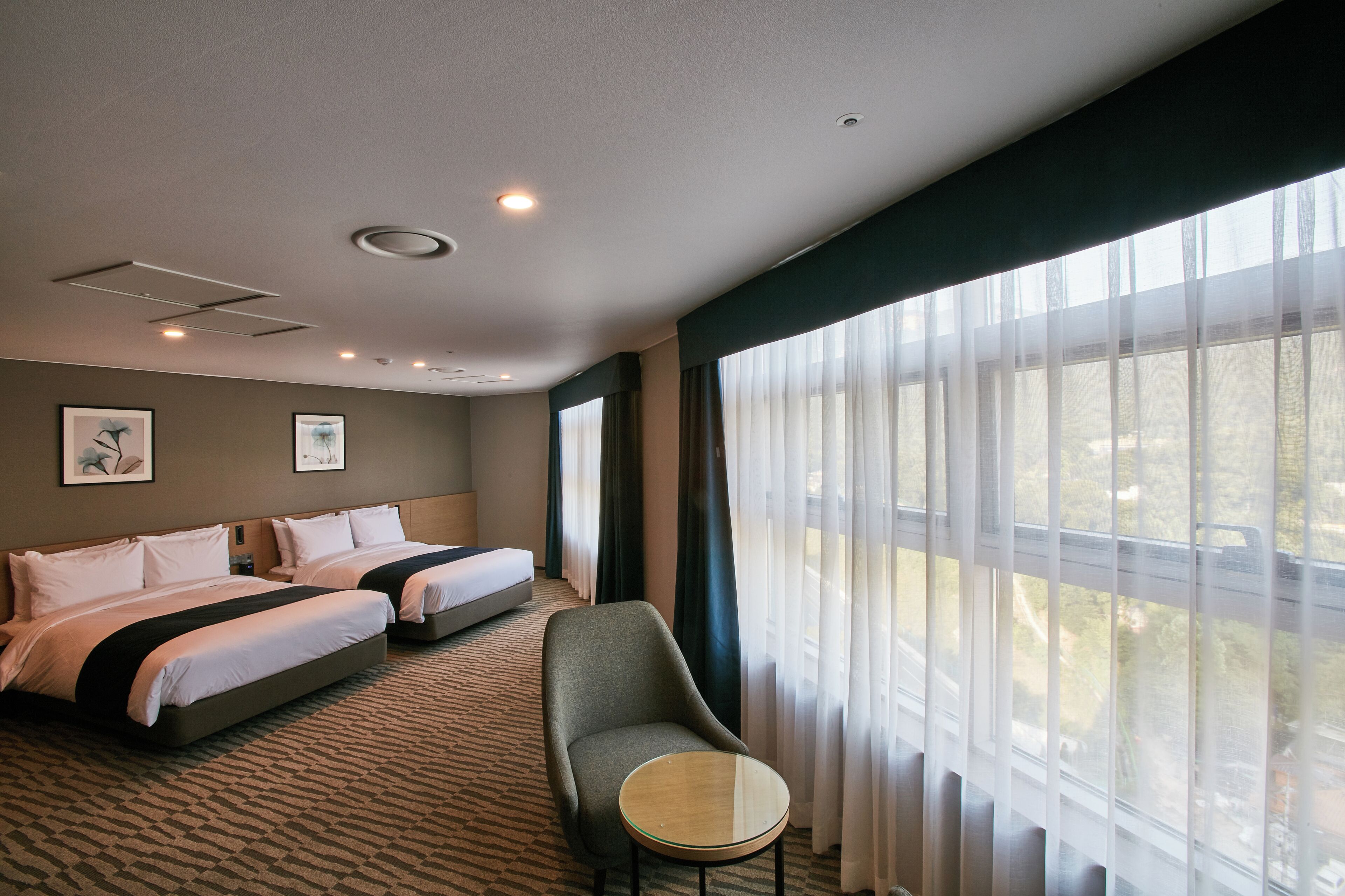 A hotel room with two beds, a chair, a small table, and a large window with curtains.
