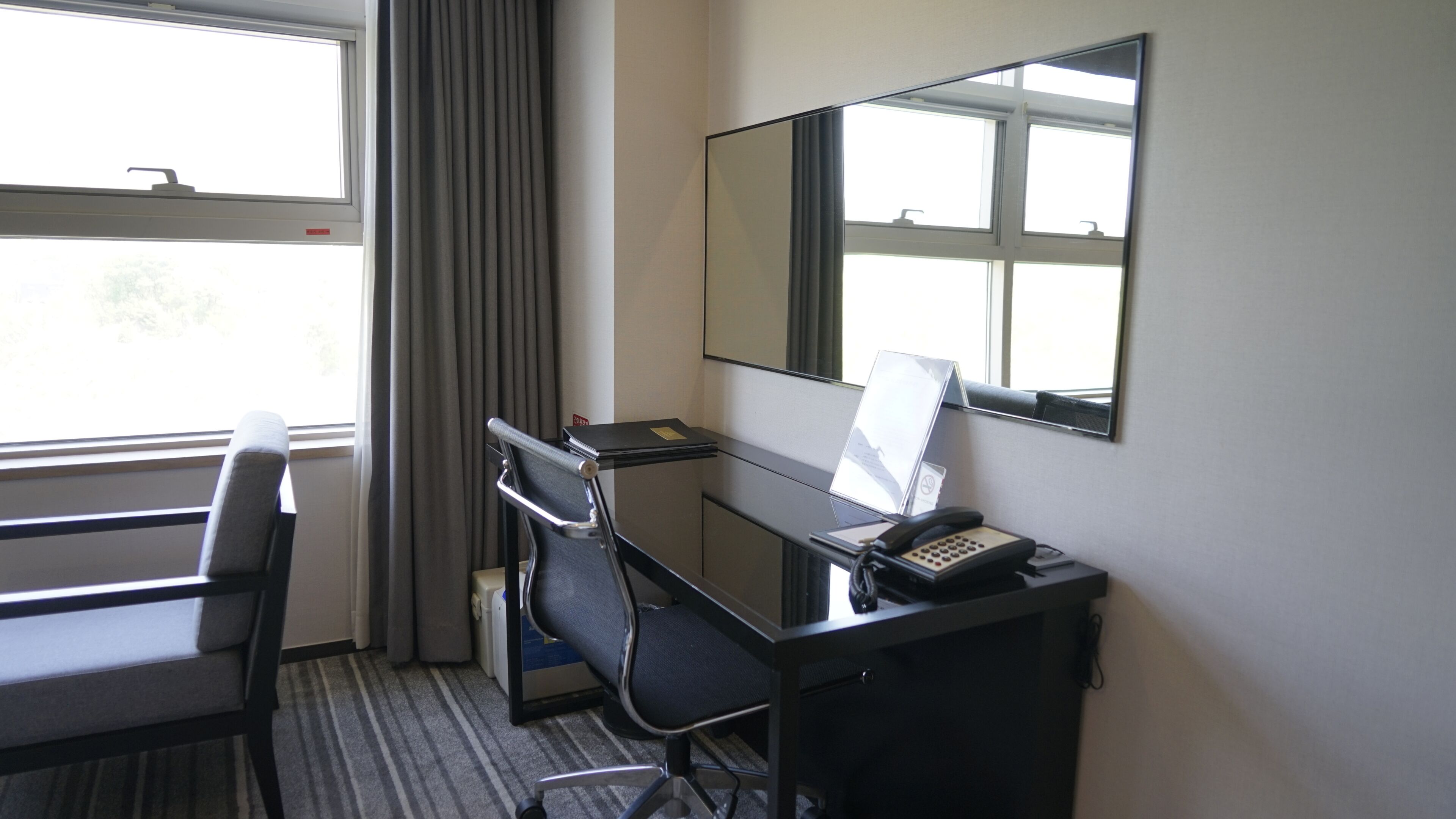 A hotel room with a desk, chair, telephone, and a large mirror.