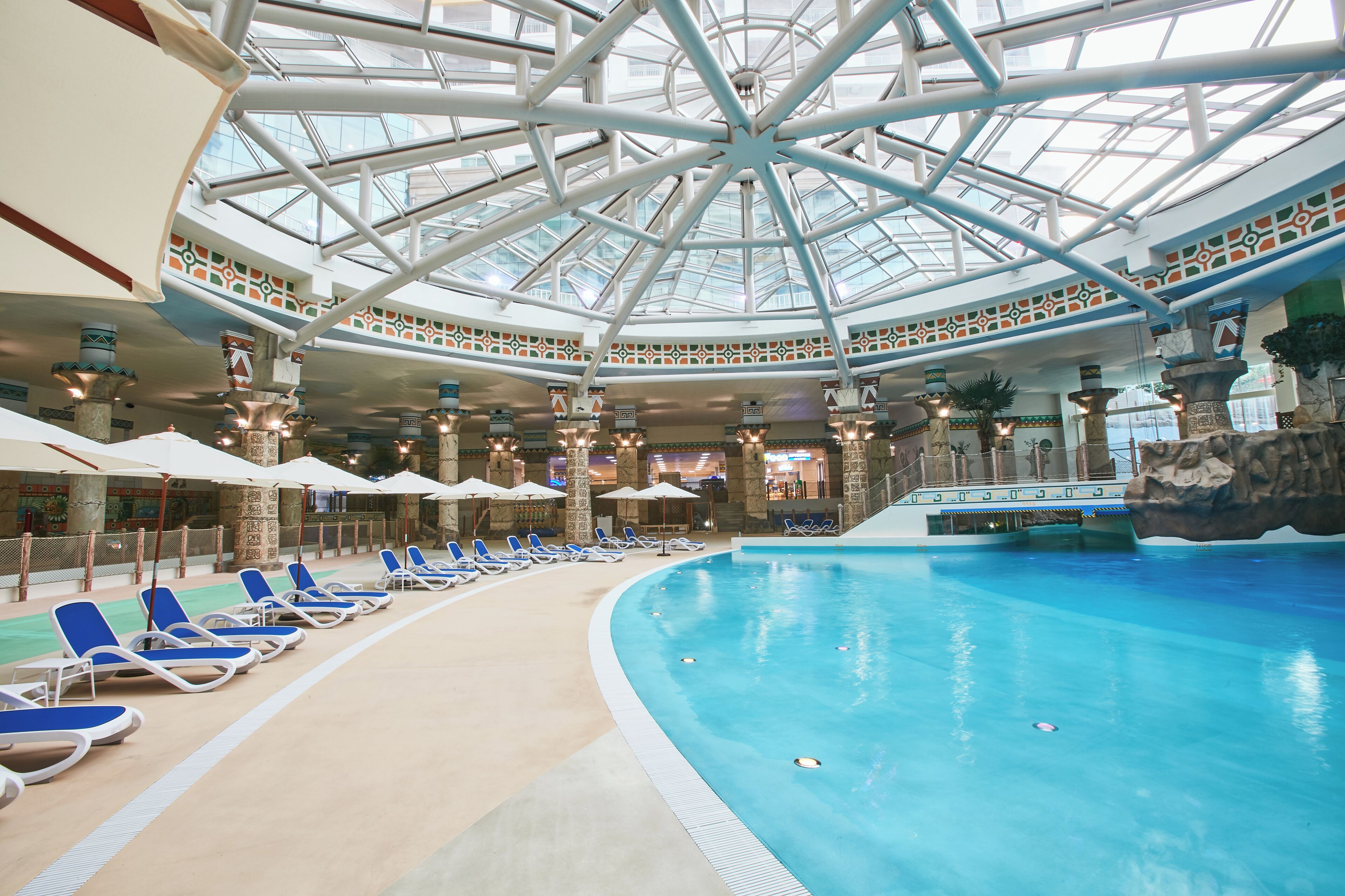 Une piscine intérieure dotée de chaises longues et de parasols, d’une verrière et de piliers décoratifs.