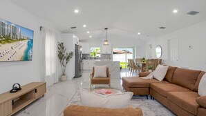 Un vaste salon comprenant un grand canapé, une table basse et un téléviseur fixé au mur. On aperçoit également un coin repas avec une table et des chaises à l’arrière-plan.