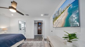 Une chambre à coucher comprenant un lit, un ventilateur de plafond, une table de chevet, un bureau, une chaise, une plante en pot, un miroir et un tableau représentant une scène de plage.