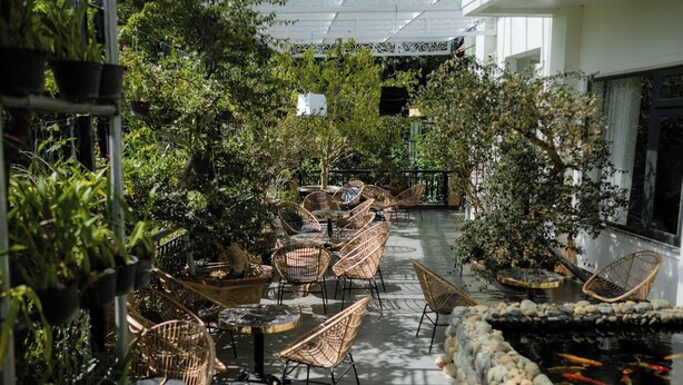 Un jardín interior con sillas y mesas de mimbre, rodeado de abundante vegetación y un estanque con peces koi.
