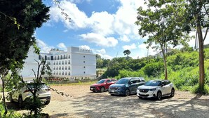 Un estacionamiento con varios coches, un edificio al fondo y vegetación circundante.