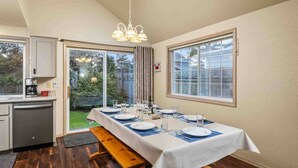 A dining area with a long table set for four, a kitchen with a coffee maker, and a view of the backyard through large sliding glass doors. - Wonderful North End Seaside Home, Fenced Backyard, Backyard Patio, 1 Mile to Shops, Food, and Fun (Seaside)