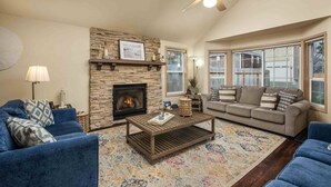 A living room with a stone fireplace, a ceiling fan, and large windows. - Wonderful North End Seaside Home, Fenced Backyard, Backyard Patio, 1 Mile to Shops, Food, and Fun (Seaside)
