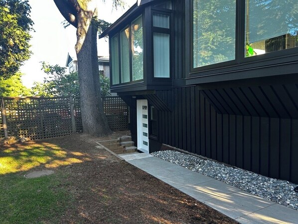 A modern two-story house with a black exterior, a visible front door, and a small front yard with a tree and a wooden fence. - 2 BDR Stylish Comfort Parkside (Vancouver)