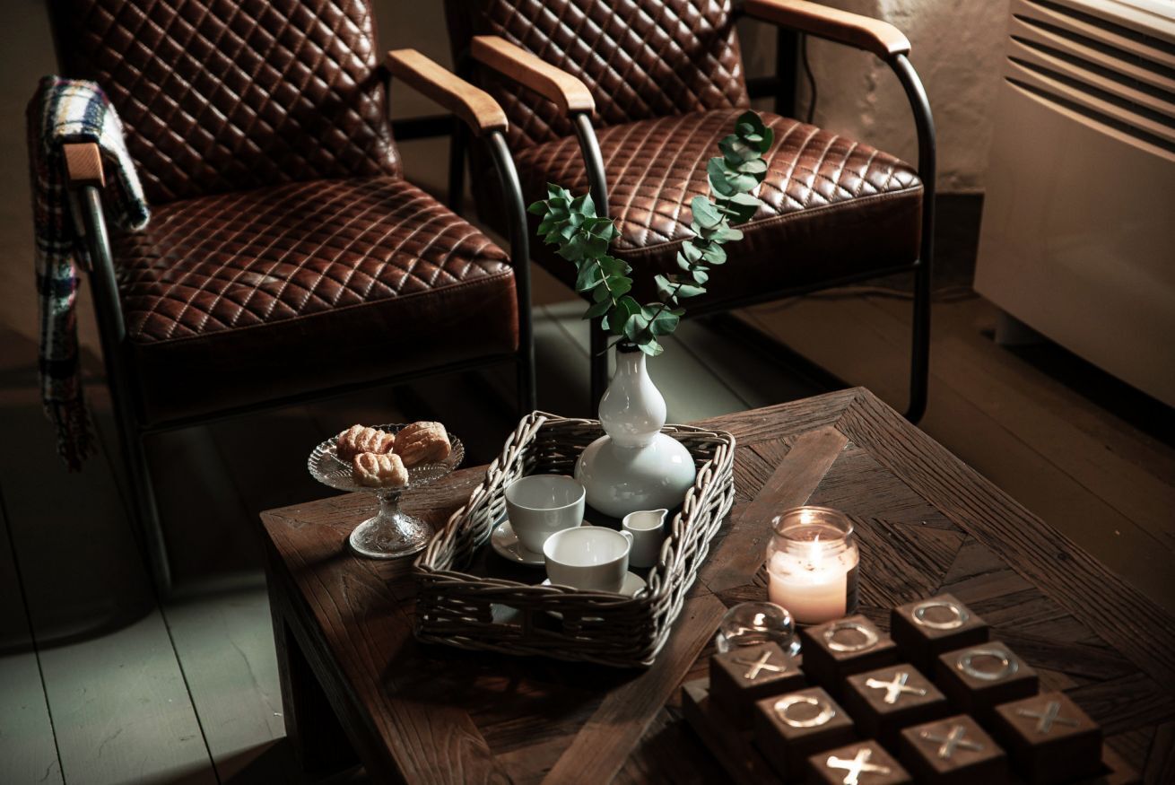 A cozy seating area with leather chairs, a wooden table with a candle, a vase with greenery, and a basket with cups and pastries.