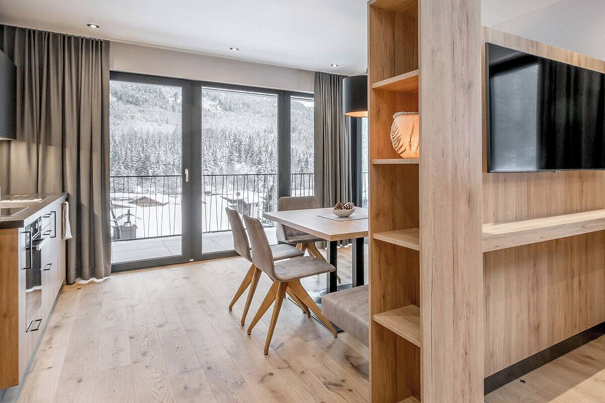 A modern living room with a flat-screen TV mounted on a wooden wall, a dining area with a table and chairs, and a view of a snowy landscape through large windows.