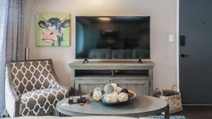 A living room with a flat-screen TV mounted above a fireplace, a patterned armchair, a coffee table with decorative balls, and a door in the background. - First Floor with 2 King Bedrooms (Kansas City)