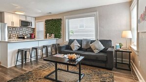 A modern living room with a sofa, coffee table, and bar stools. A kitchen area with a microwave and stove is visible in the background. - Cute Midtown 1 Bedroom (Kansas City)