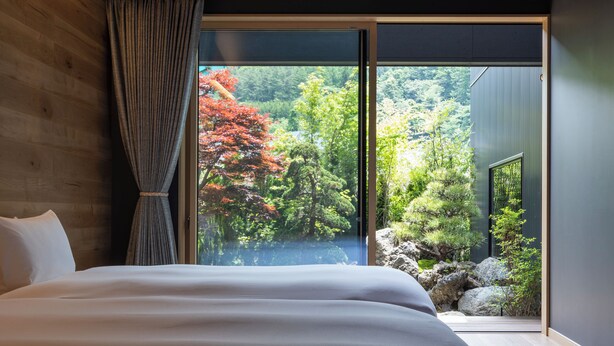 Une chambre dotée d’un grand lit, donnant sur un jardin avec des arbres et des rochers, et possédant une porte-fenêtre coulissante qui mène à un espace extérieur.