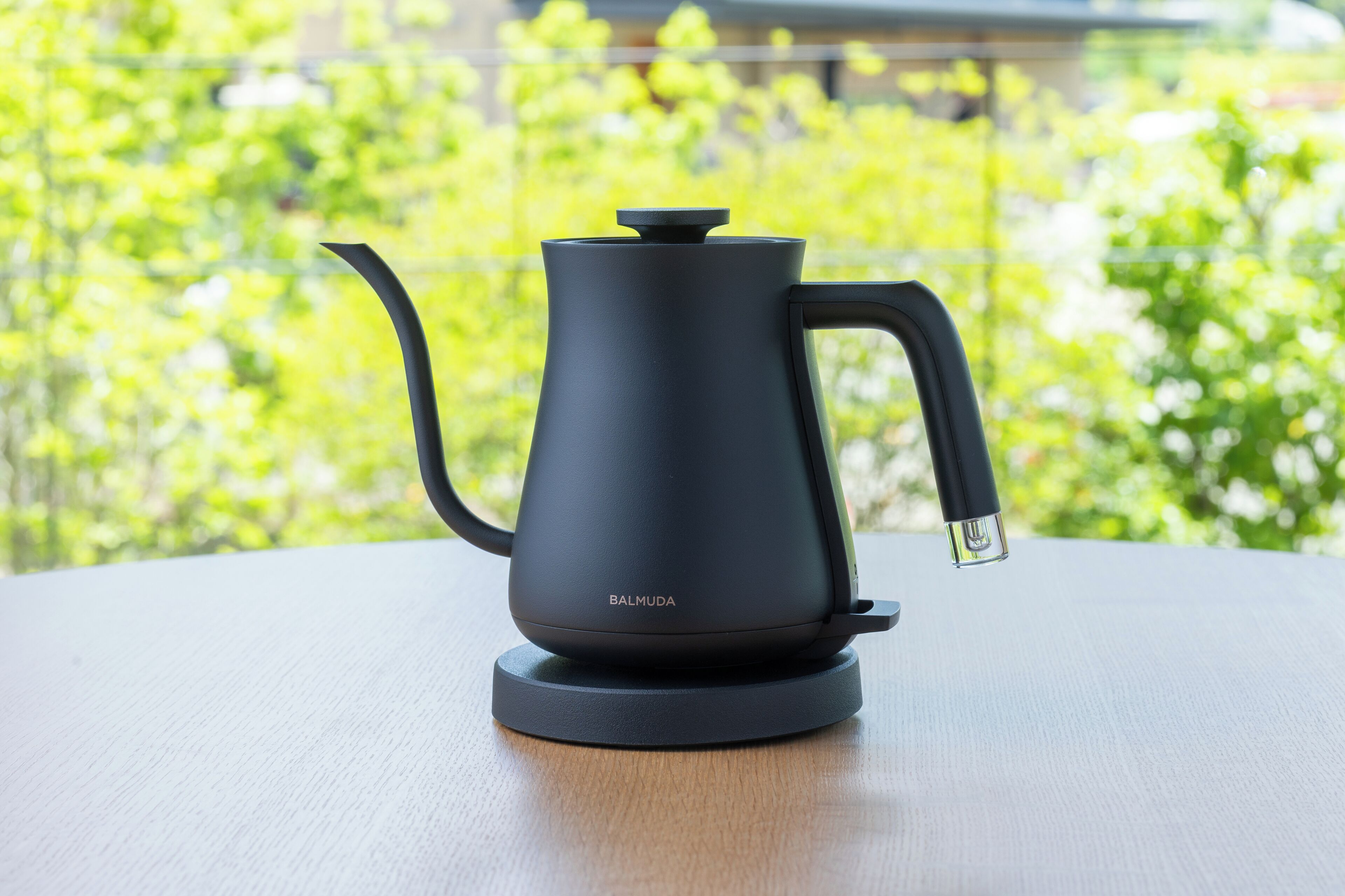 A black Barmuda coffee maker on a wooden table with a green, leafy background.
